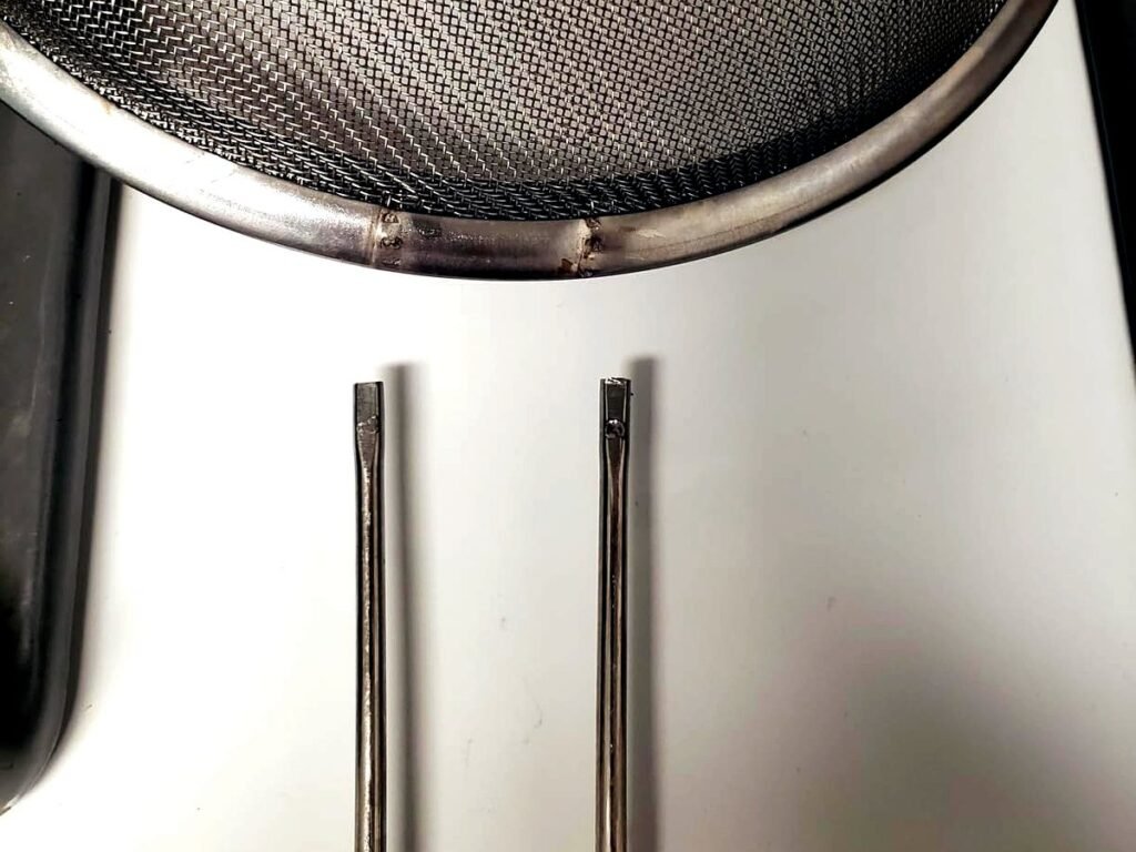 Close-up of a broken strainer rim showing the two failed spot weld marks and the snapped ends of the wire handle